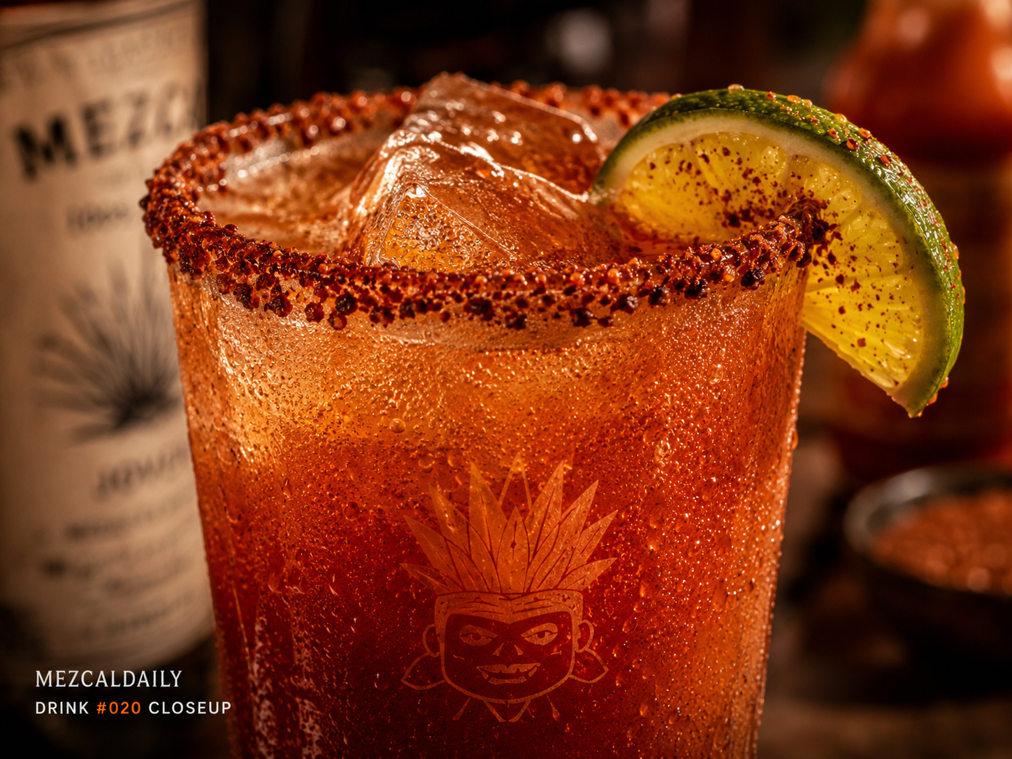 Close-up of the Mezcal Michelada with chile-salt rim, red savory beer cocktail, ice, lime wedge, and condensation.